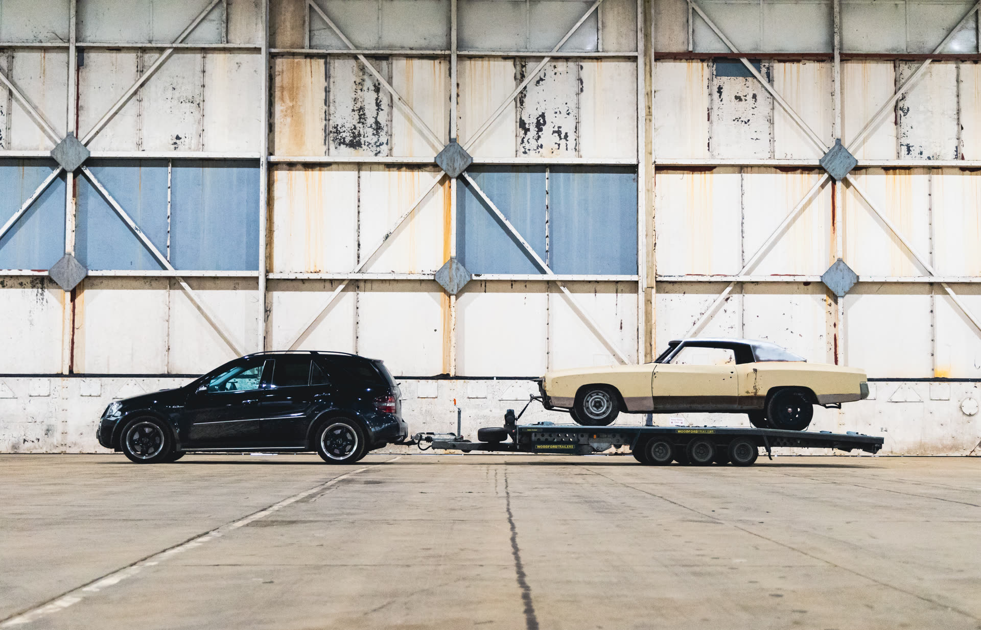 A Mercedes ML towing a classic American coupe on a trailer outside a workshop hangar